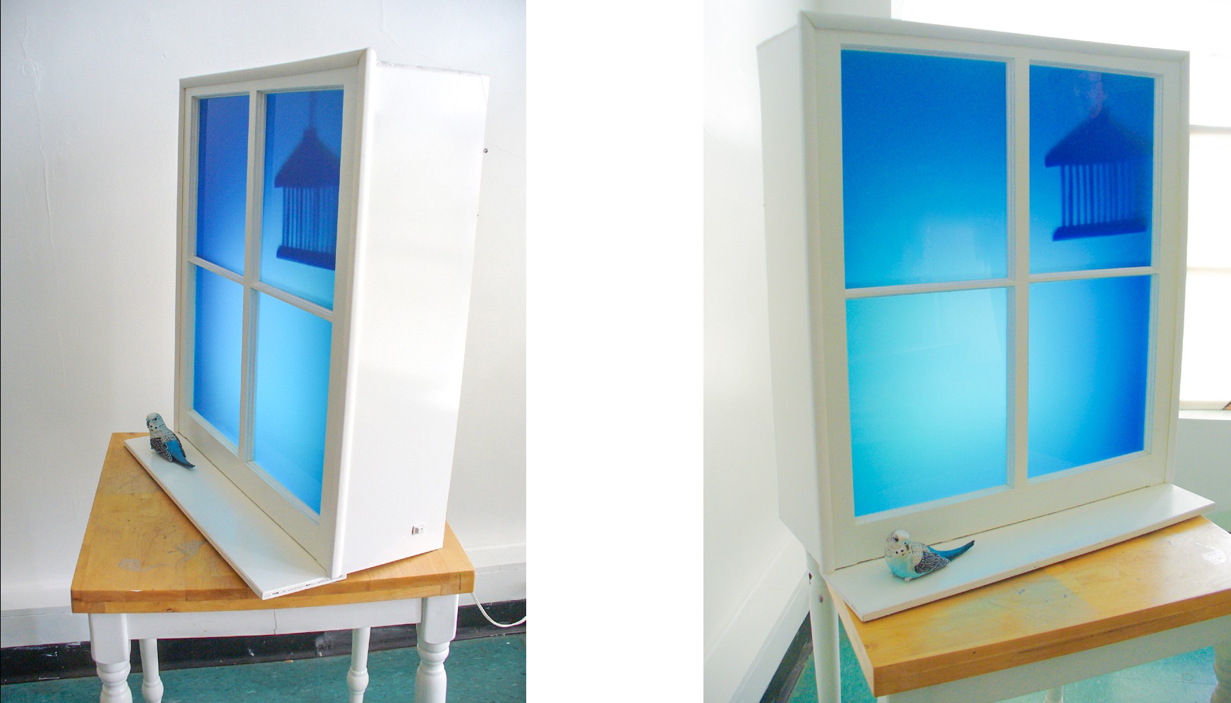 A freestanding window rests on a table. It is a light box that looks like a window, and the panes are glowing blue. A small bird sculpture rests on the ledge.
