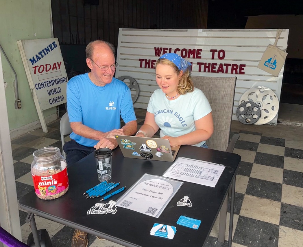 A man and woman sit at a table in front of a sign that says welcome to the theatre.