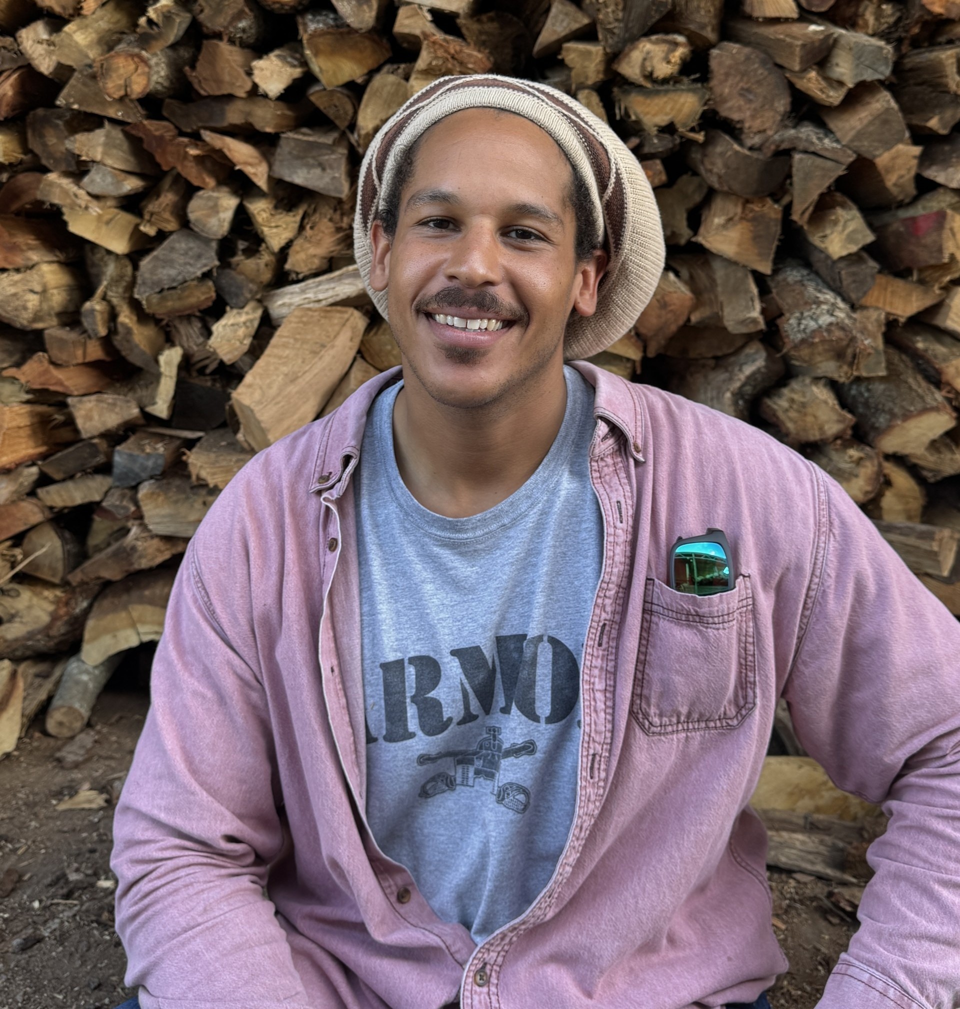 A man in a hat poses with a pile of wood logs.
