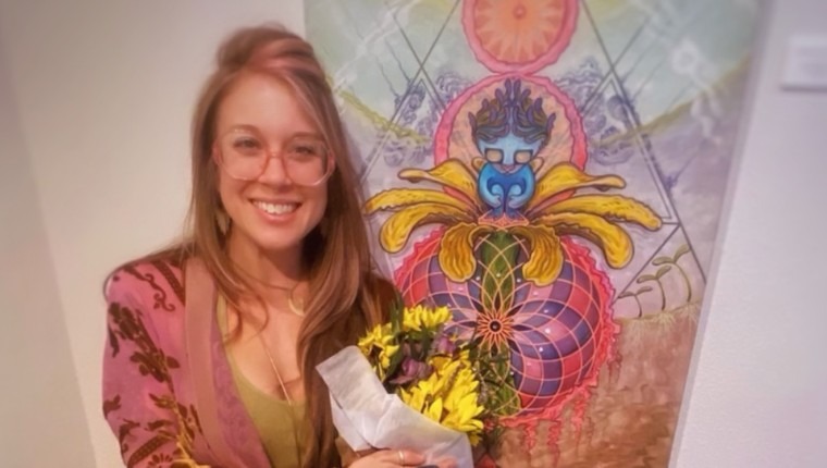 A woman smiles and holds flowers in front of a painting.